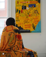 Load image into Gallery viewer, Mercedes Cook wrapped in Hollywood Dreams blanket by Andromeda Cook featuring Basquiat-inspired design, 528hz frequency, Self Portrait as a Legend, M31 Andromeda galaxy, and divine feminine energy with original painting in background