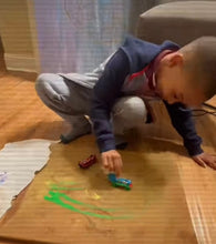 Load image into Gallery viewer, Young artist Lamonte creating Tracks of Imagination by dipping toy trains in green paint and driving them across cardboard to form abstract lines and movement